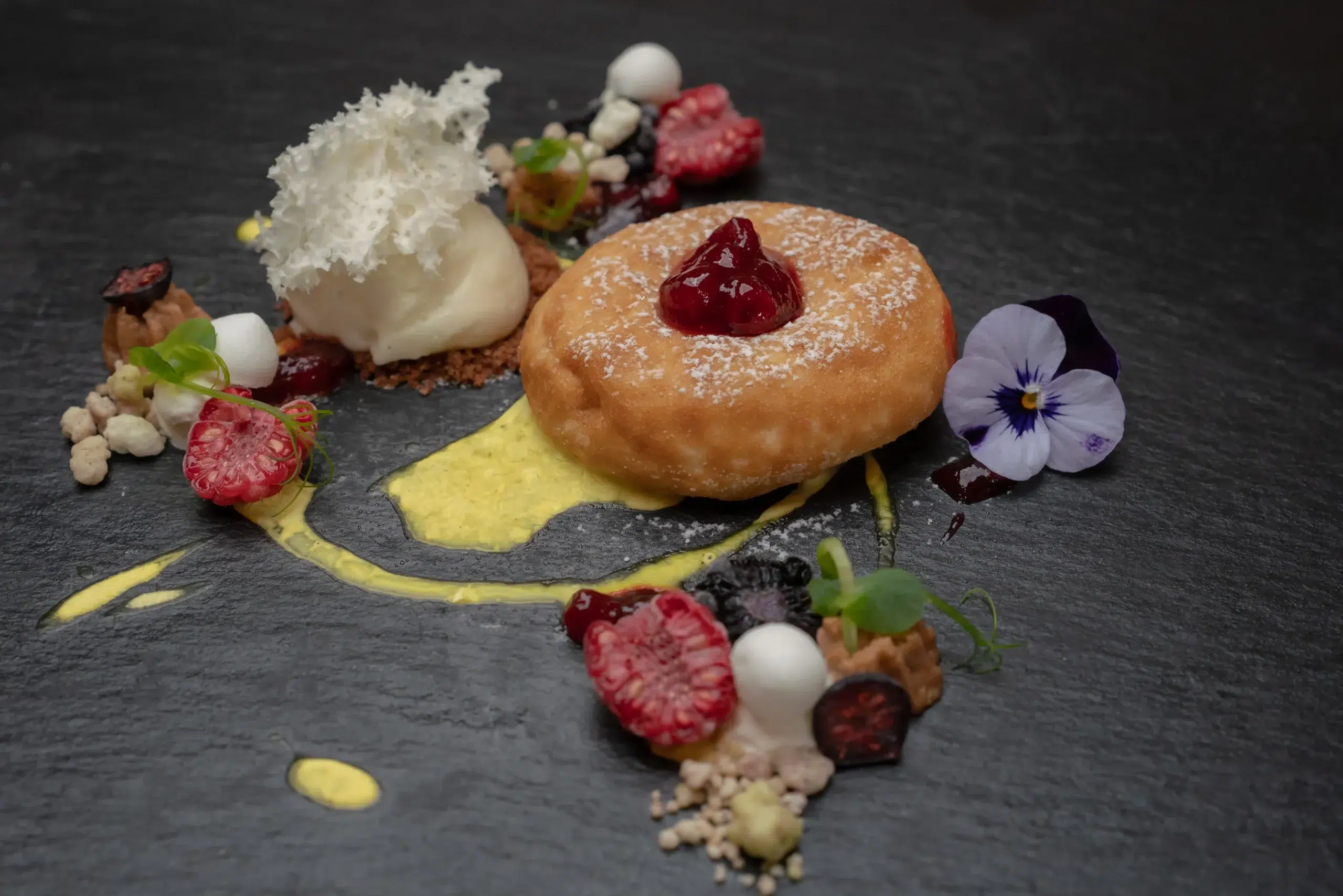 Auf einem Teller serviertes Restaurantdessert mit Donut, Eiscreme, Himbeeren, essbaren Blüten und Soße.
