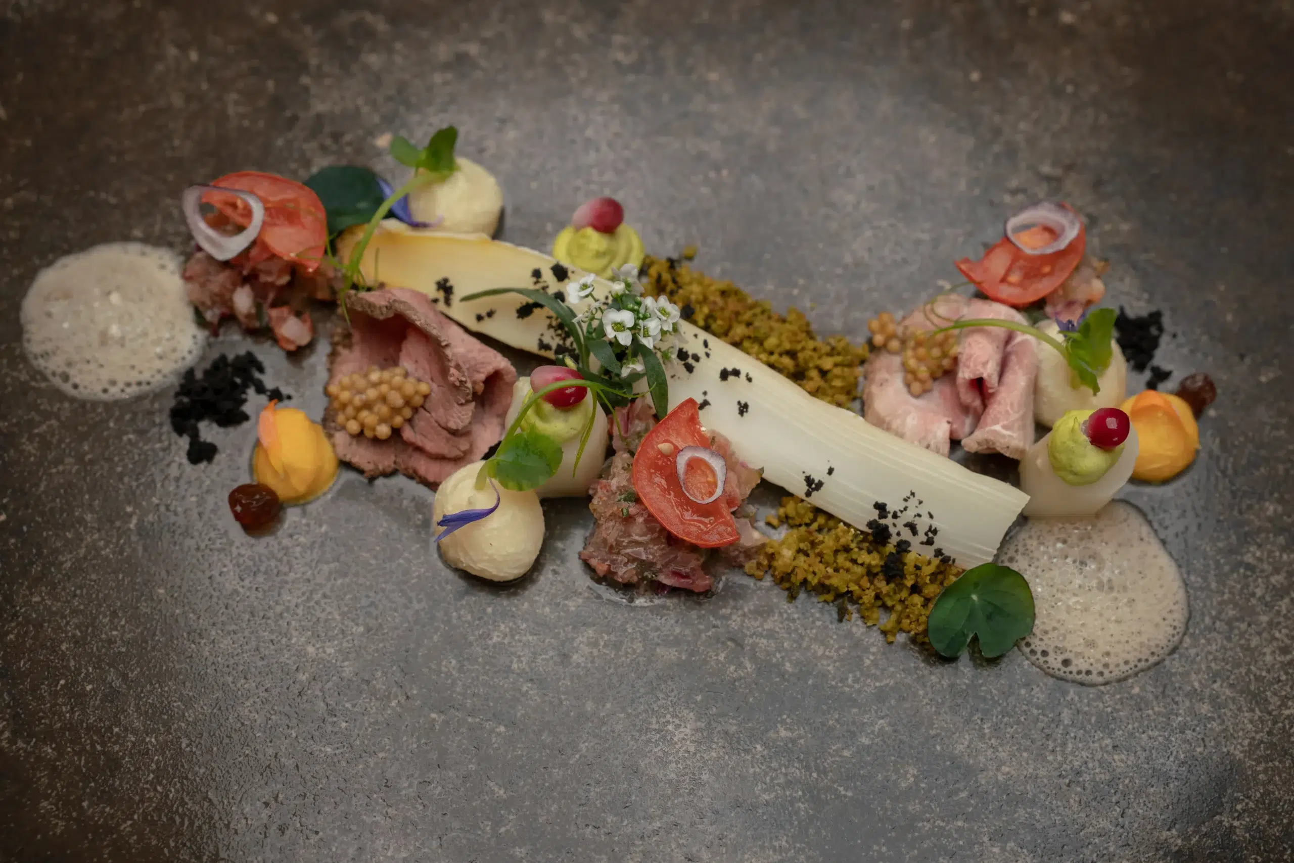 Gourmet-Restaurantgericht mit Fleisch, Soßen, Garnituren und Schaum, kunstvoll auf einem dunklen Teller angerichtet.