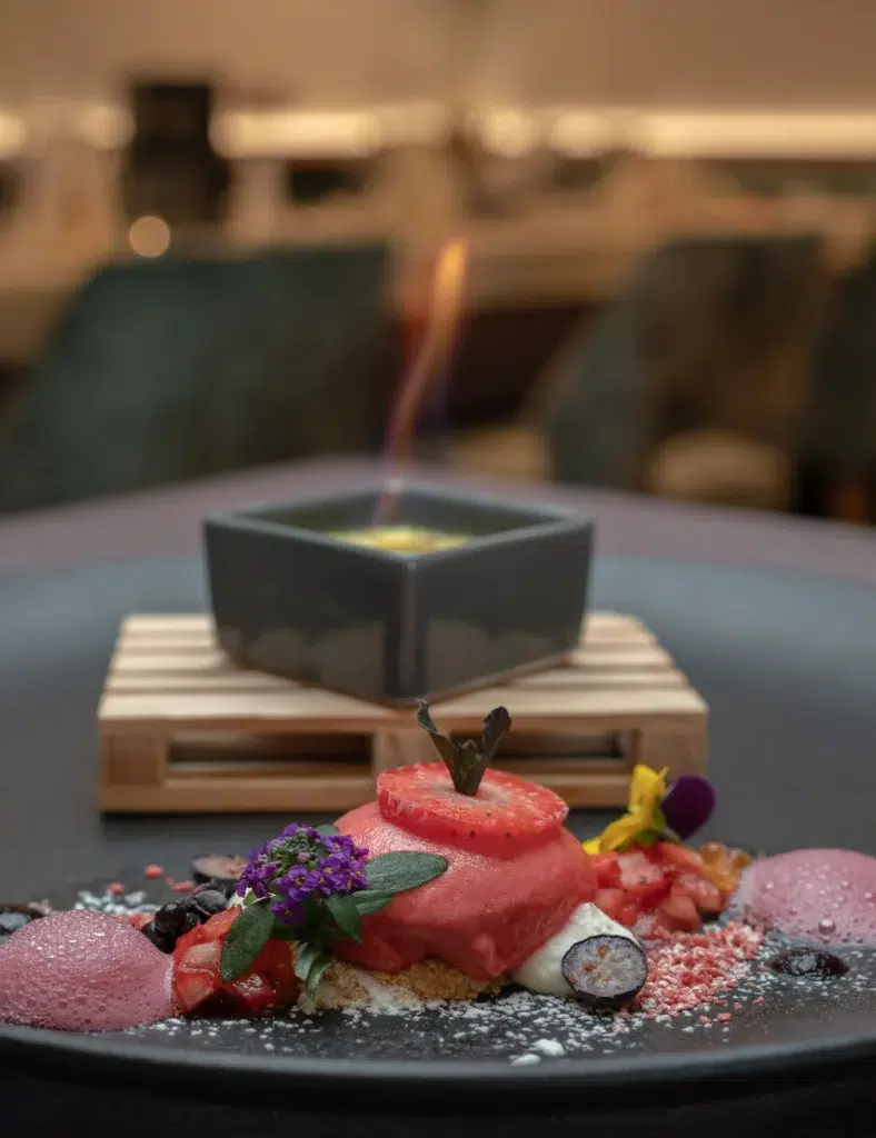 Gourmet-Restaurant-Dessert mit Beeren, Blumen und einer kleinen brennenden quadratischen Schale im Hintergrund.