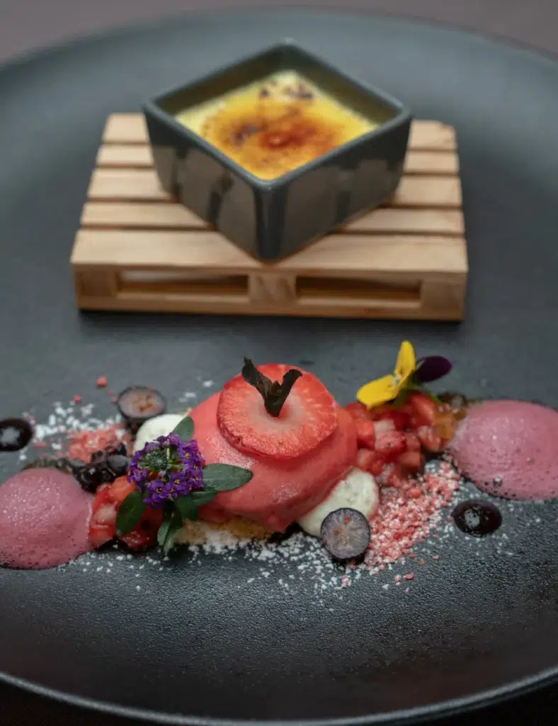 Gourmet-Dessert im Restaurantstil mit Beeren und Blumen, serviert mit Crème Brûlée auf einem schwarzen Teller.