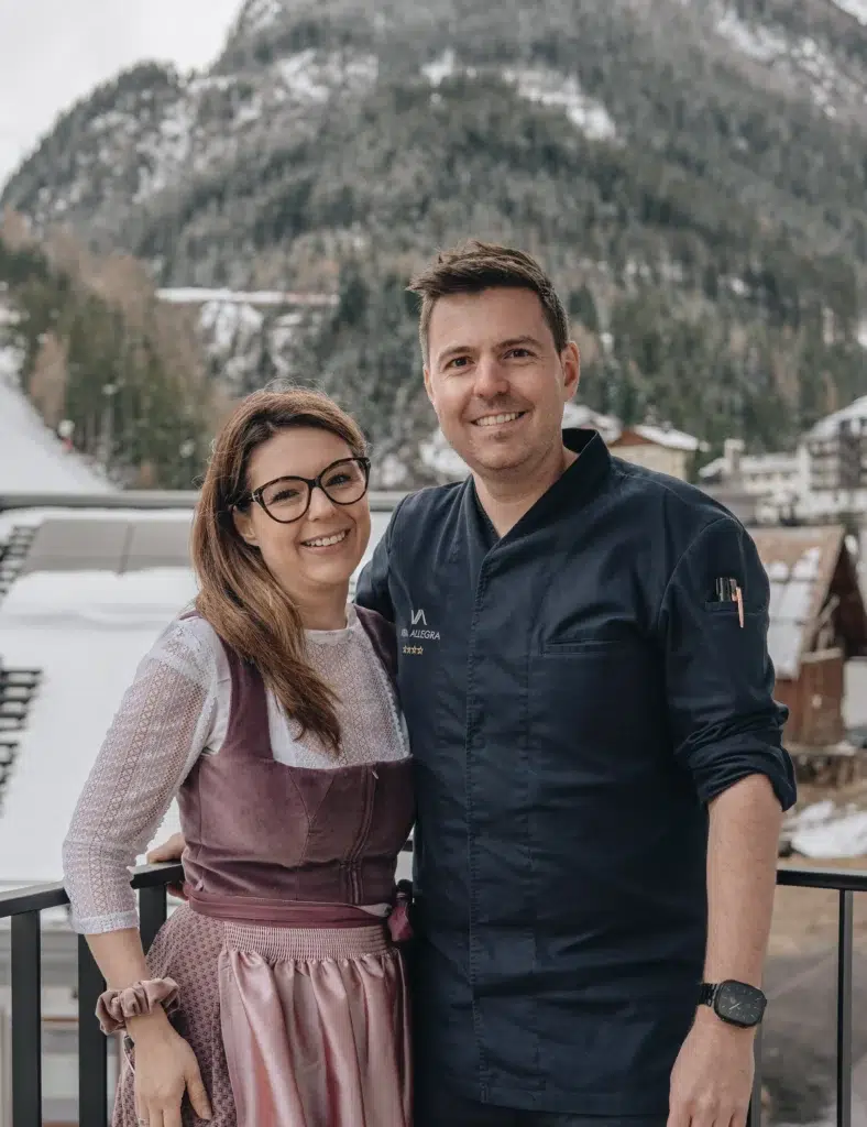 Ein lächelndes Paar steht auf einem Vista Allegra-Balkon mit schneebedeckten Bergen und Hütten im Hintergrund.