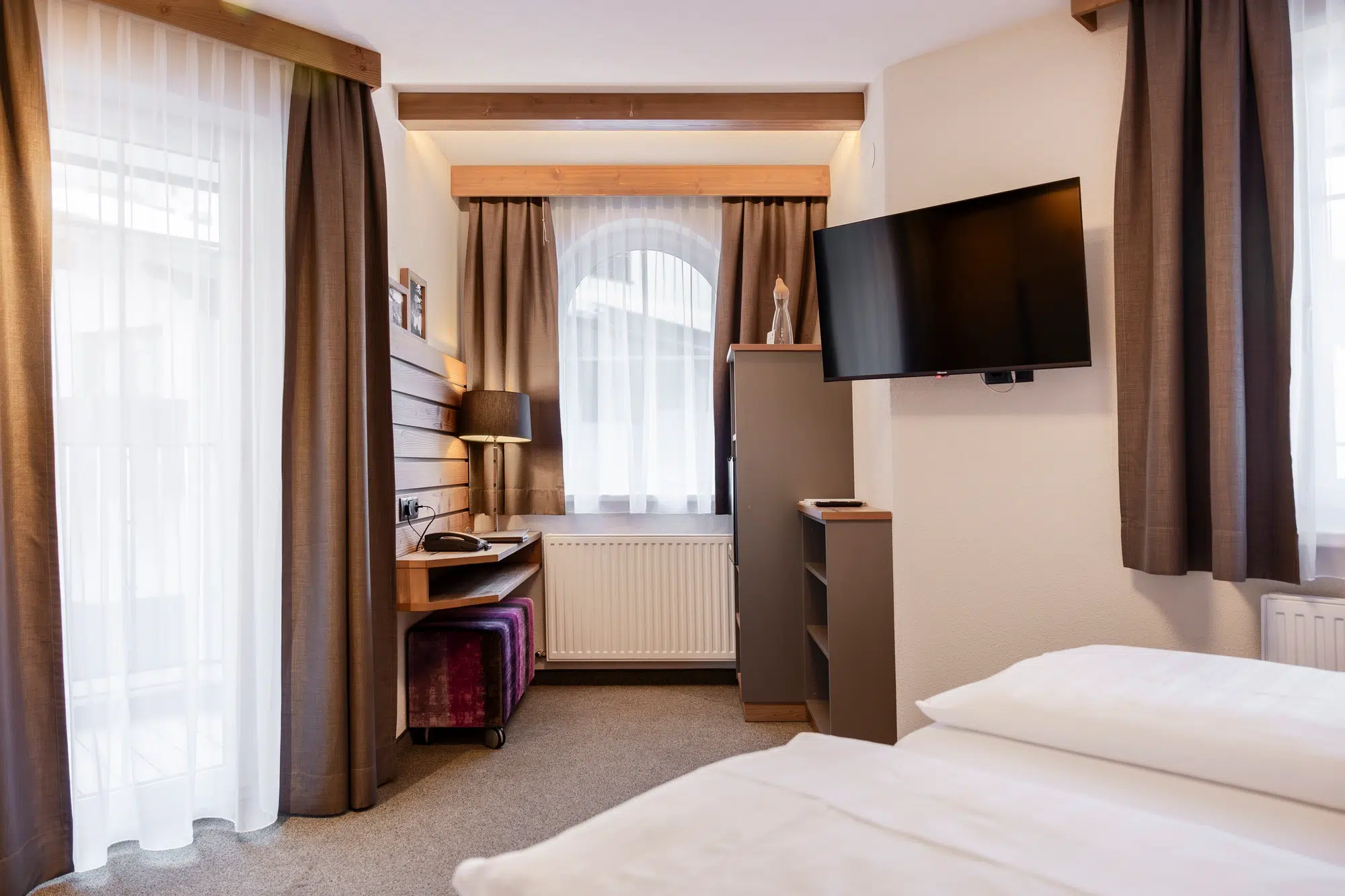 Modernes Zimmer mit Bett, Schreibtisch, Fernseher und großen Fenstern mit braunen Vorhängen.