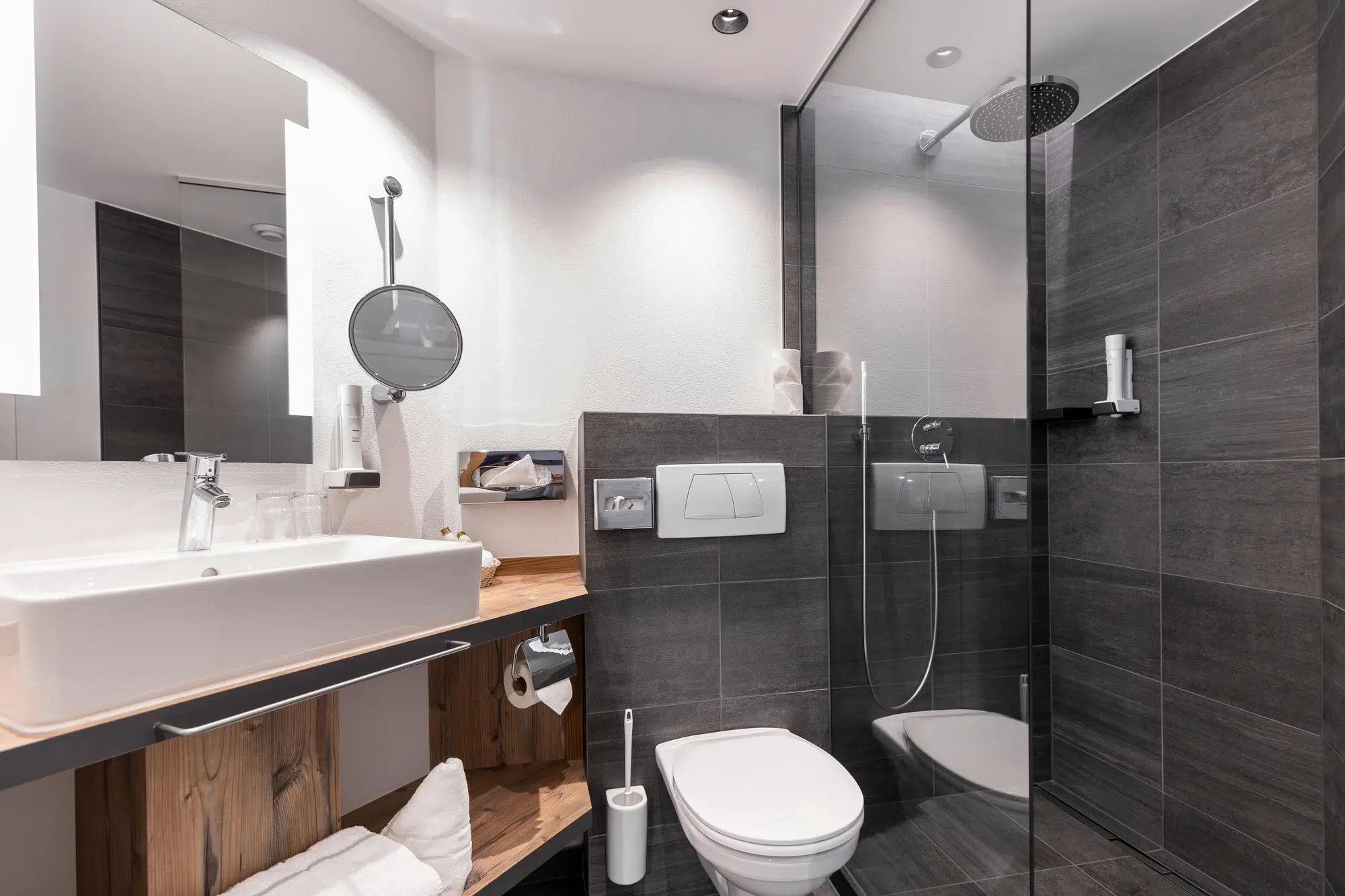 Modernes Badezimmer Zimmer mit ebenerdiger Dusche, Toilette, Waschbecken und dunkelgrau gefliesten Wänden.