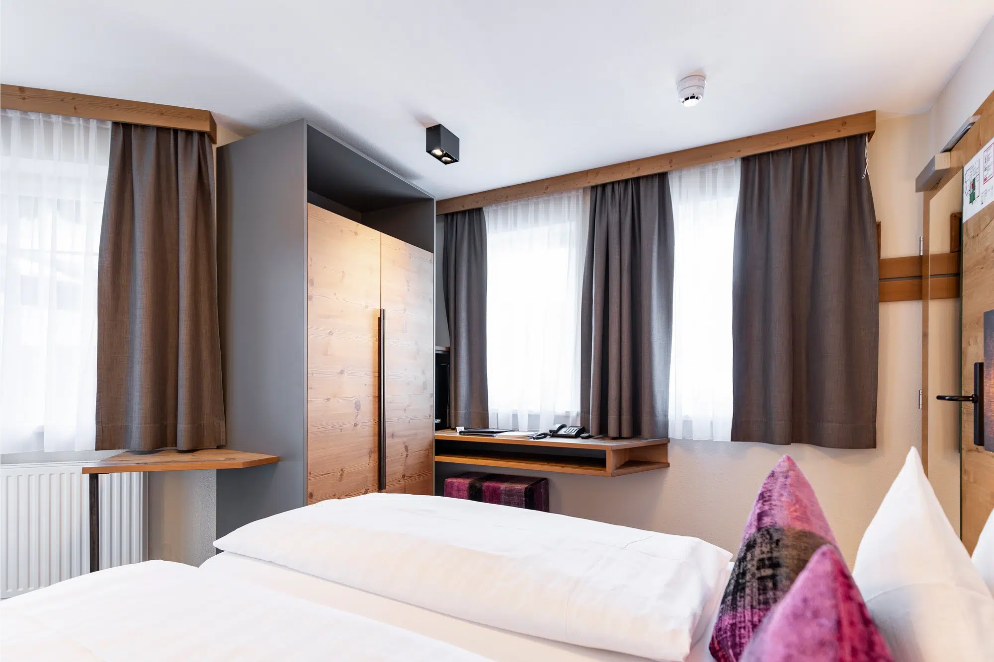 Modernes Zimmer mit Bett, Schreibtisch, Kleiderschrank und großen Fenstern mit grauen Vorhängen.