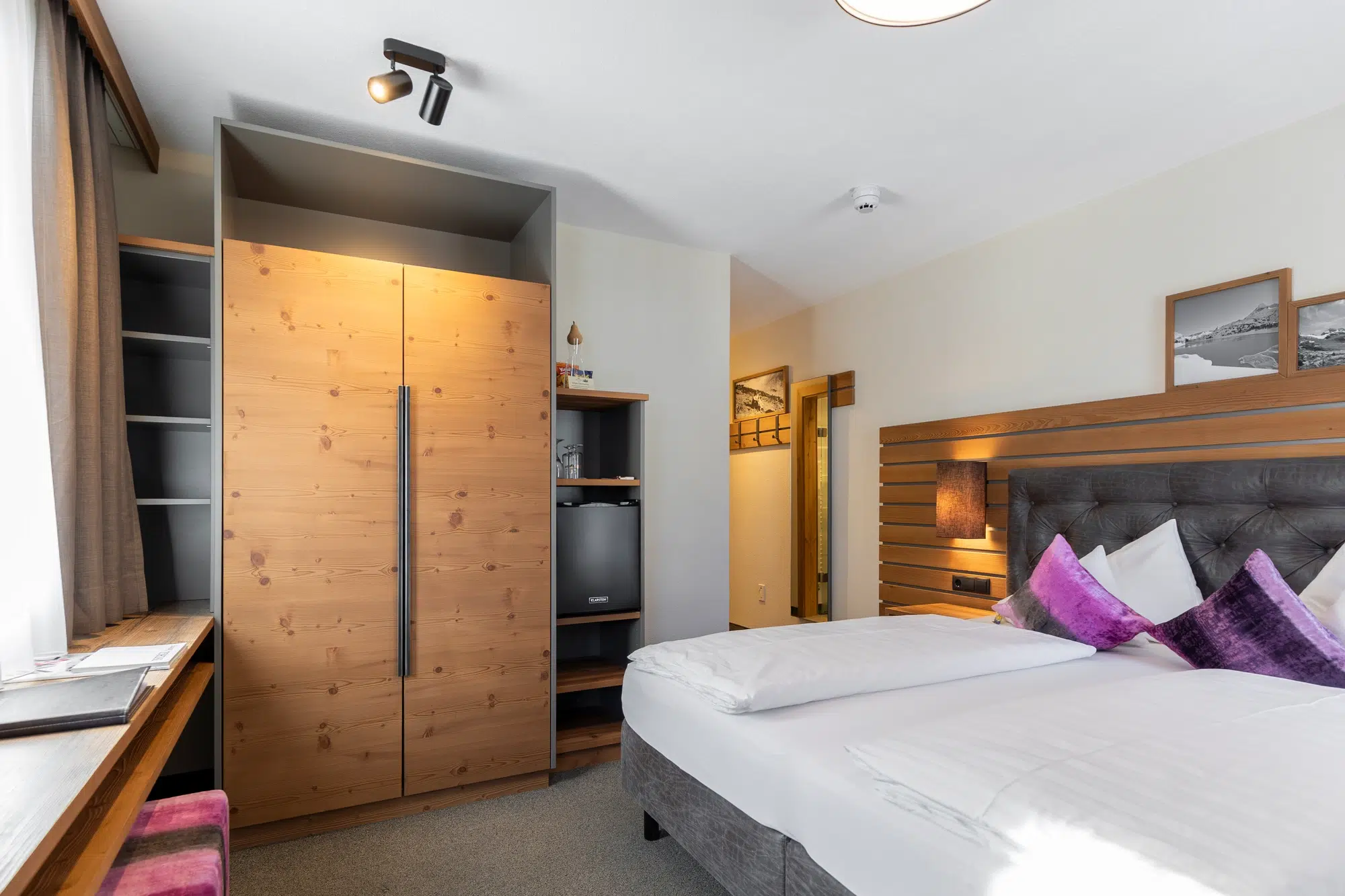 Zimmer mit zwei Einzelbetten, Holzschrank, Schreibtisch und stilvollen violetten Akzentkissen.
