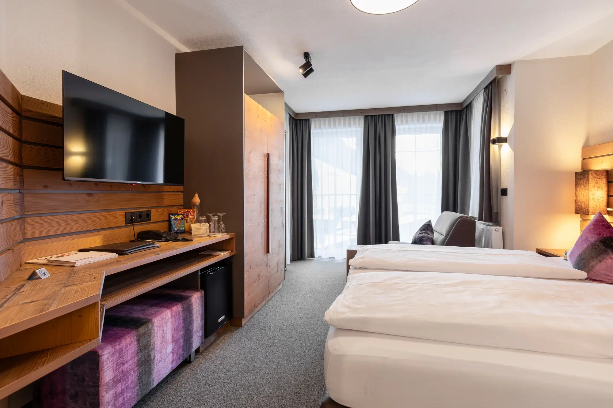 Modernes Zimmer mit zwei Einzelbetten, Fernseher, Schreibtisch, Minibar und einem großen Fenster mit Vorhängen.