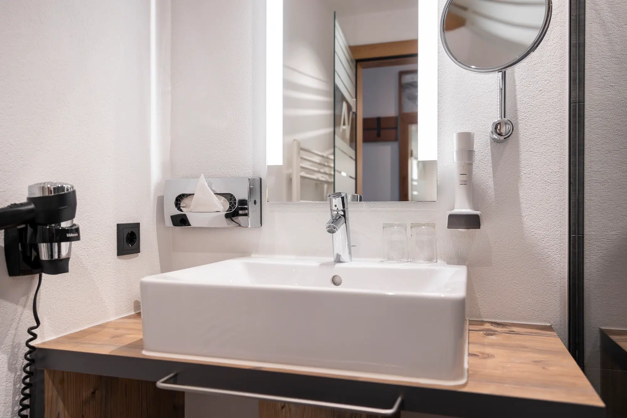Modernes Waschbecken im Zimmer-Badezimmer mit Spiegel, Brille, Taschentuchbox und Haartrockner auf Holzplatte.