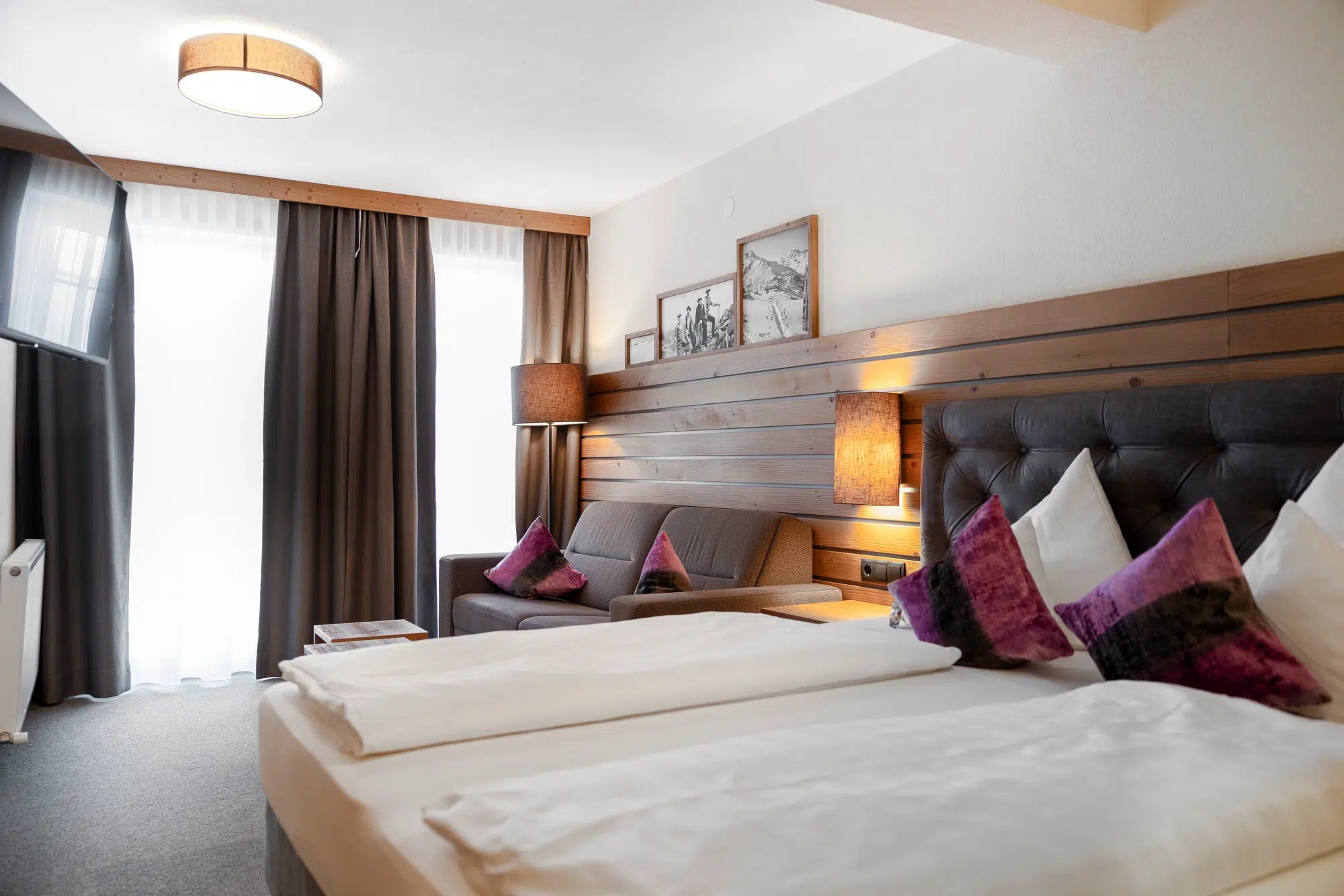Modernes Zimmer mit Doppelbett, Sofa und großem Fenster mit dunklen Vorhängen.