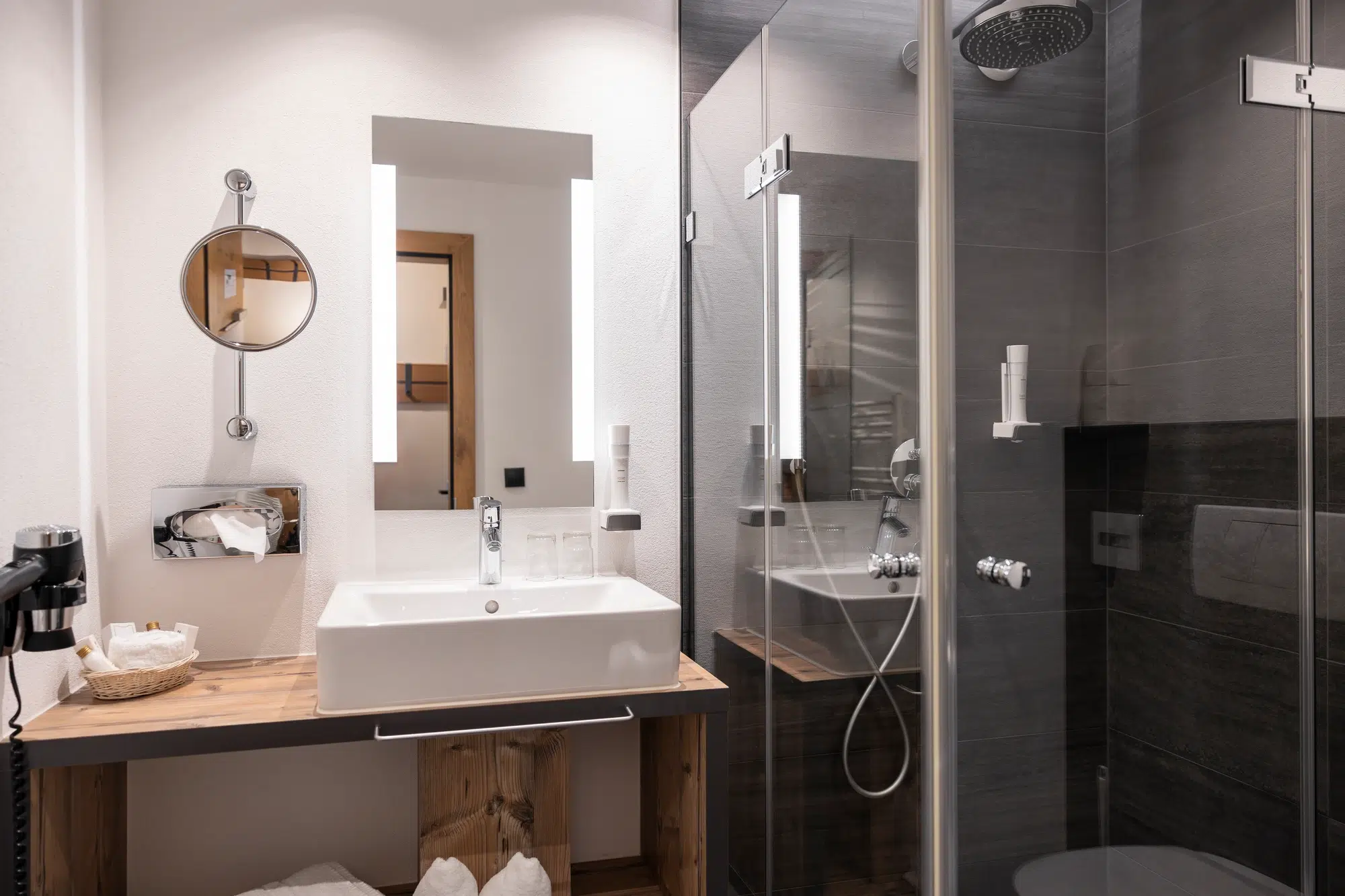 Modernes Zimmer-Badezimmer mit Waschbecken, rundem Spiegel, Glasdusche und Holzarbeitsplatte.