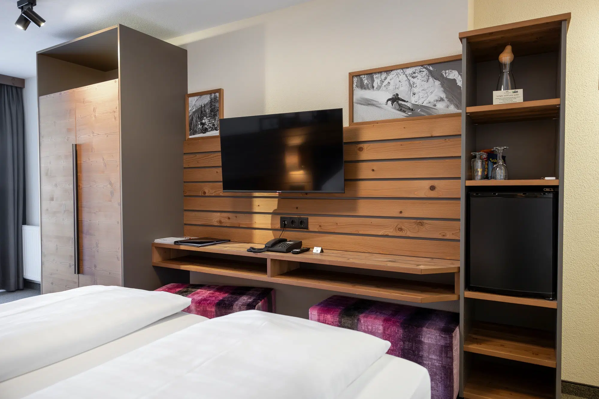 Modernes Zimmer mit Holzakzenten, Fernseher, Kühlschrank und zwei Betten im Vordergrund.