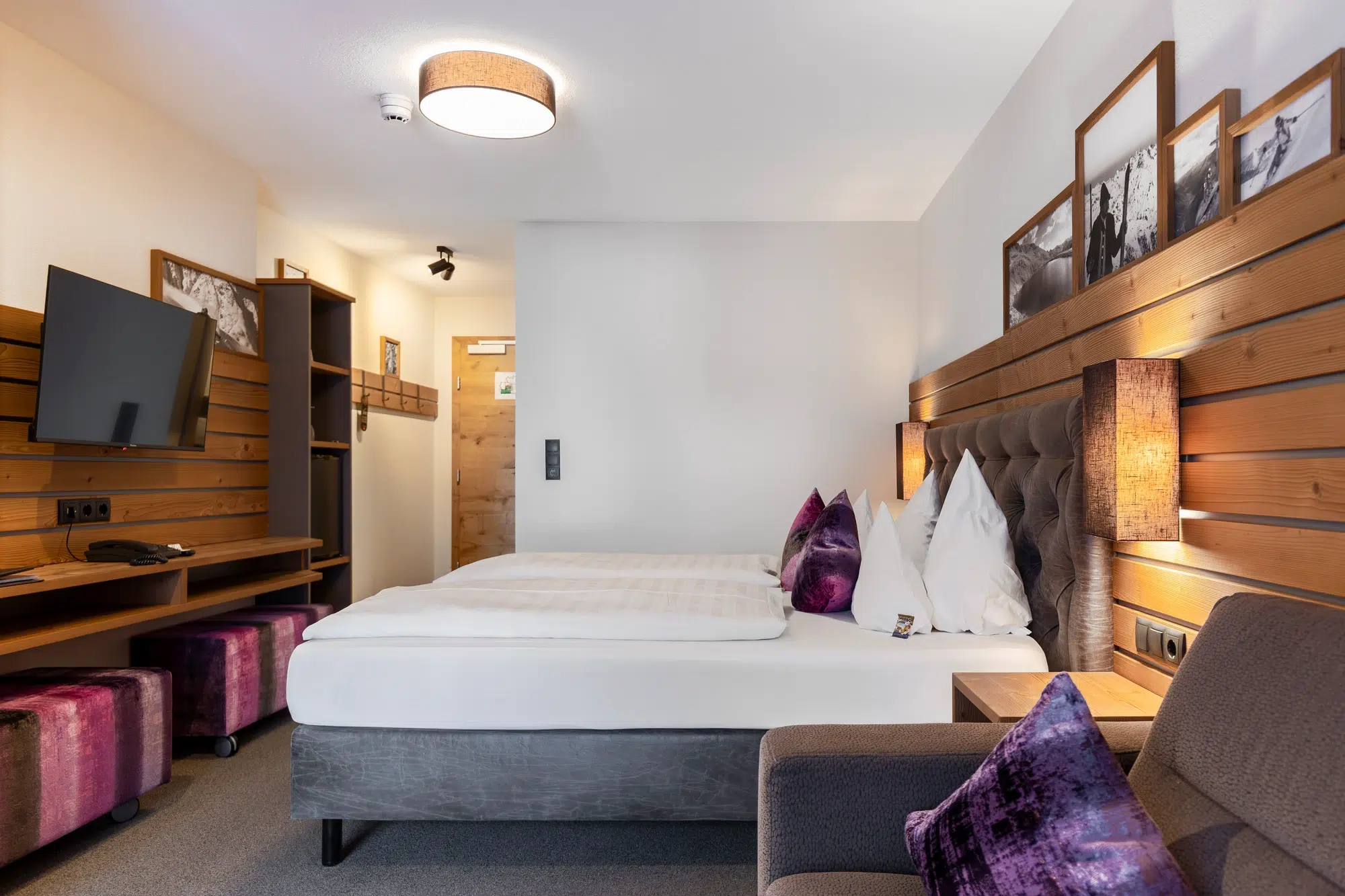 Modernes Zimmer mit Doppelbett, Fernseher, Holzakzenten und stilvollen lila Kissen.