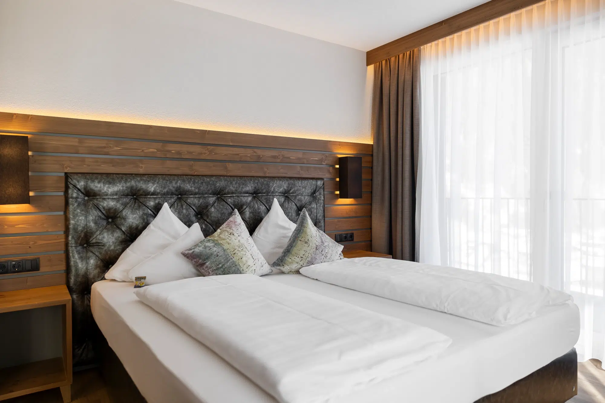 Modernes Zimmer mit weichem Bett, Holzakzenten und großem Fenster mit eleganten Vorhängen.