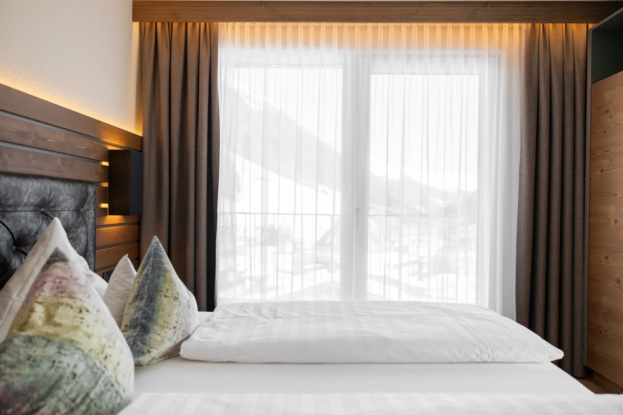 Ein ordentlich gemachtes Zimmer-Bett neben einem Fenster mit durchsichtigen Vorhängen und Blick auf die Berge draußen.