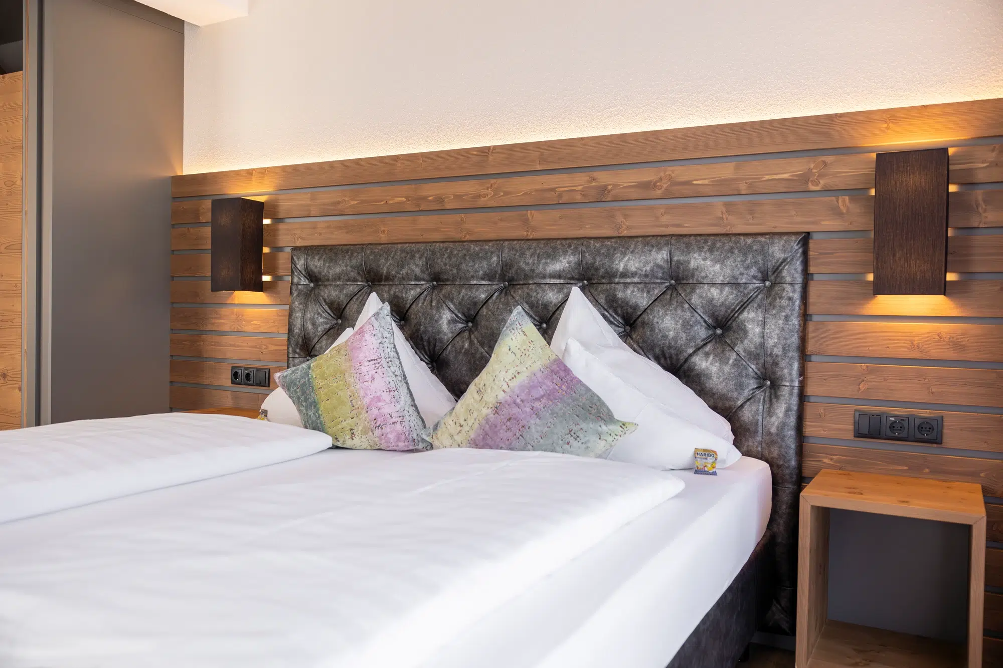 Modernes Zimmer mit Doppelbett, bunten Kissen, Holzkopfteil und Wandleuchten.