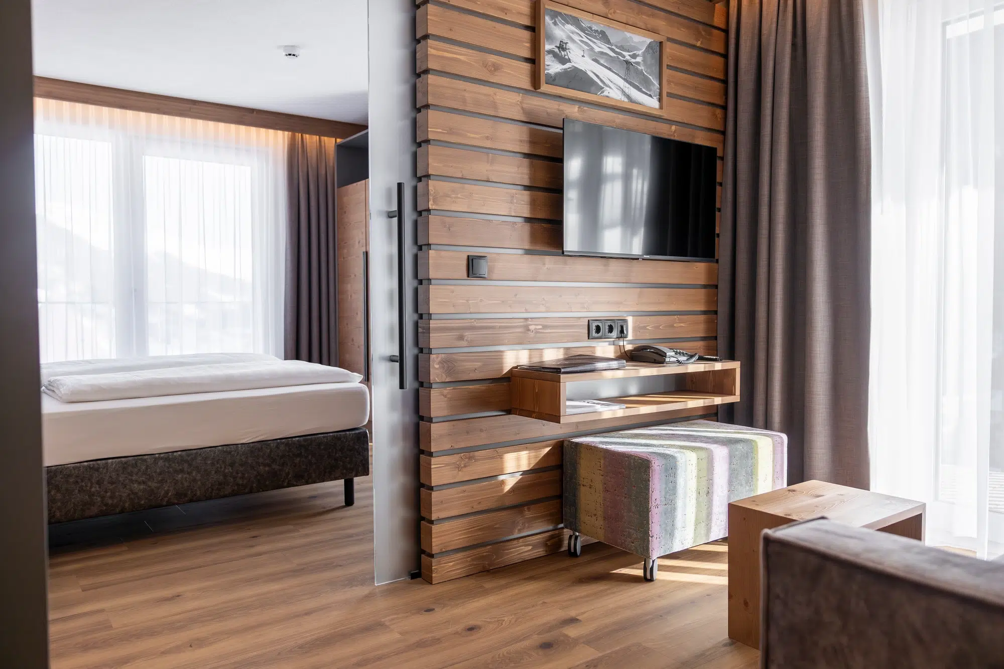 Modernes Zimmer mit Holzakzenten, gemütlichem Bett, an der Wand montiertem Fernseher und einer bunten Bank.