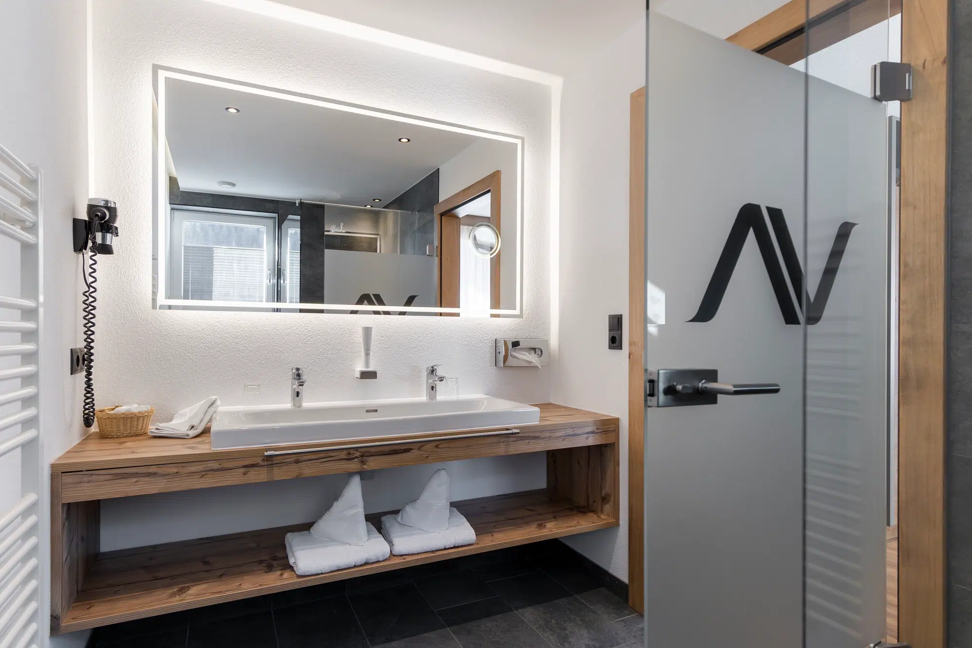 Modernes Badezimmer Zimmer mit großem Spiegel, Doppelwaschbecken, Holzregal und Milchglastür.