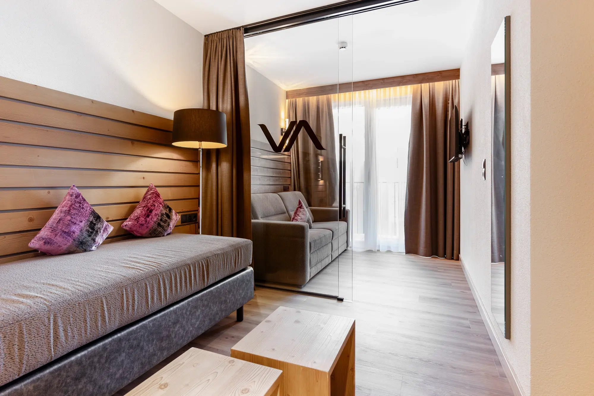 Modernes Zimmer mit Bett, Sofa, Glastrennwand und raumhohen Vorhängen.