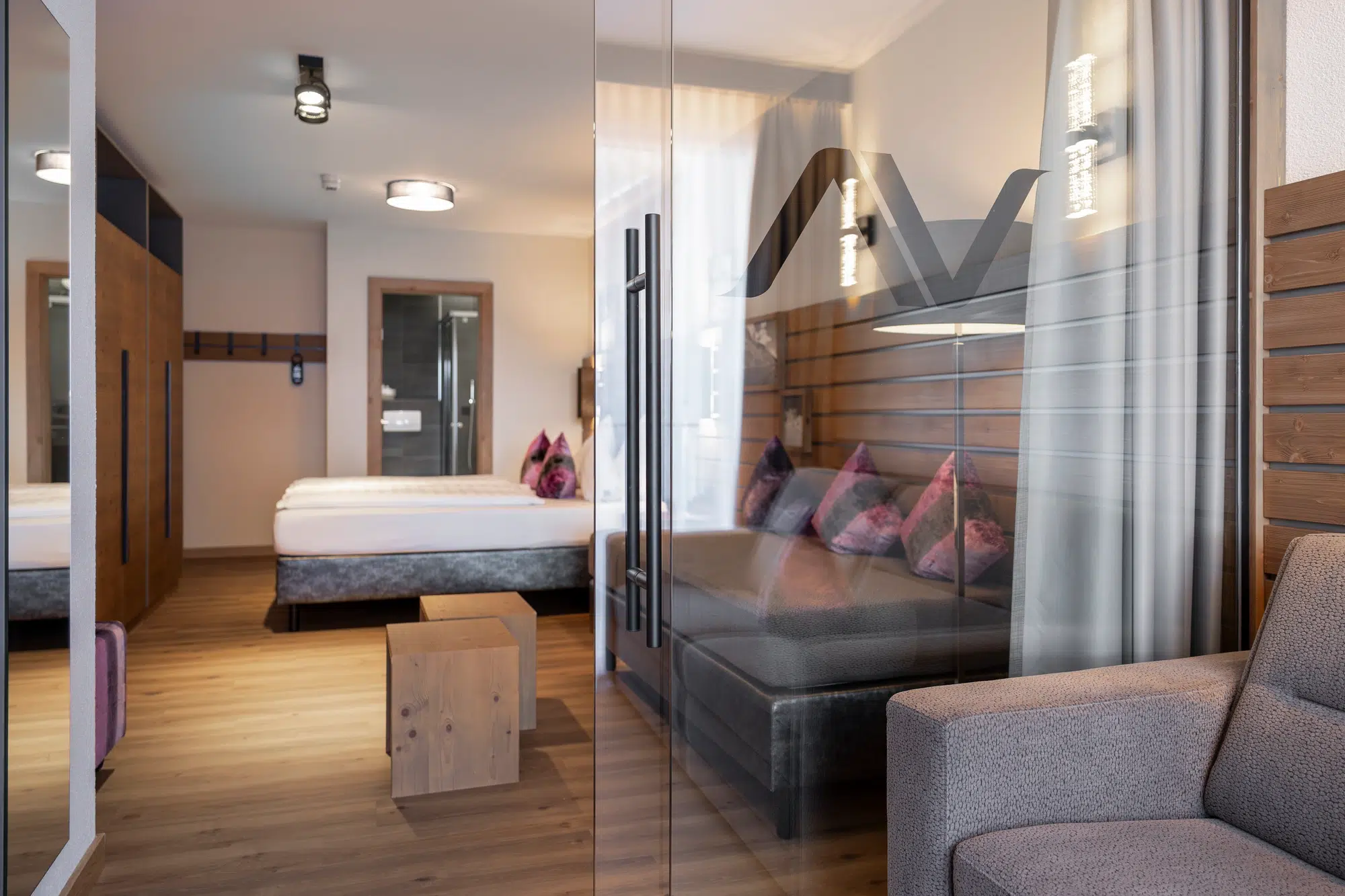 Modernes Zimmer mit gemütlichem Bett, Sofa und Holzakzenten, durch eine Glastrennwand sichtbar.