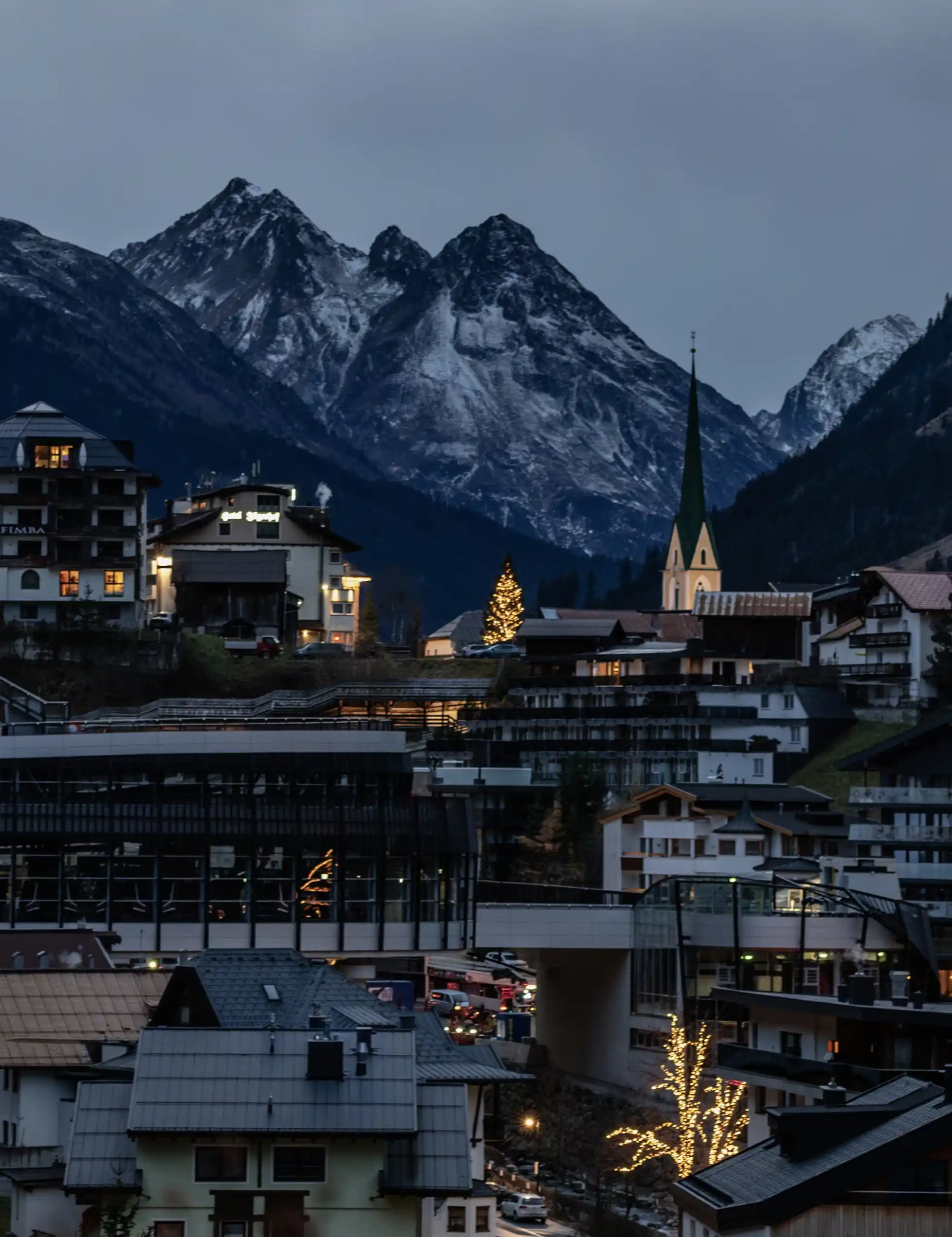 Ein Bergdorf in der Abenddämmerung mit beleuchteten Gebäuden, einem Kirchturm, schneebedeckten Gipfeln, Lage & Anreise.