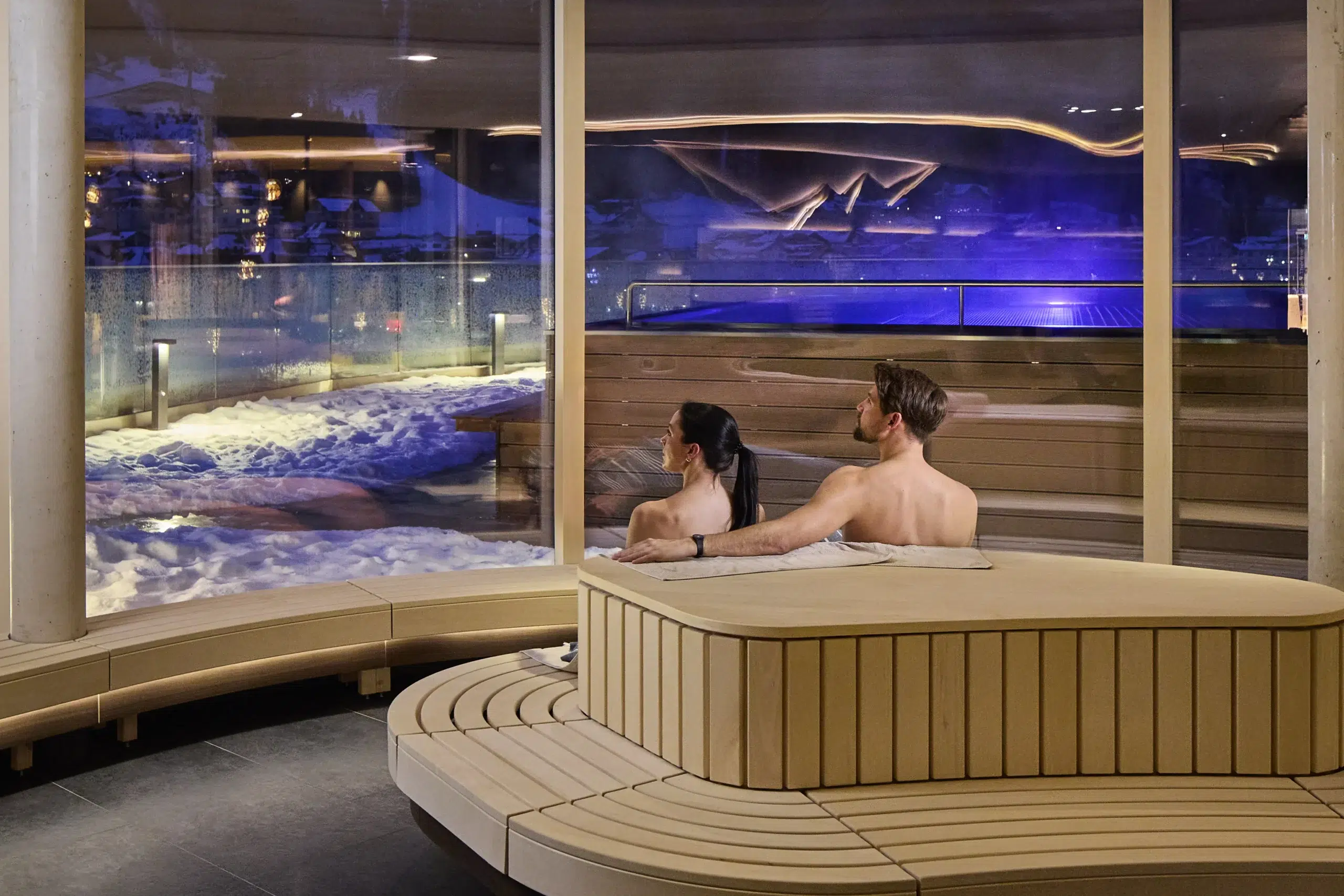 Ein Paar entspannt sich in einem Whirlpool im Innenbereich eines Wellnesshotels und blickt auf die verschneite Landschaft.