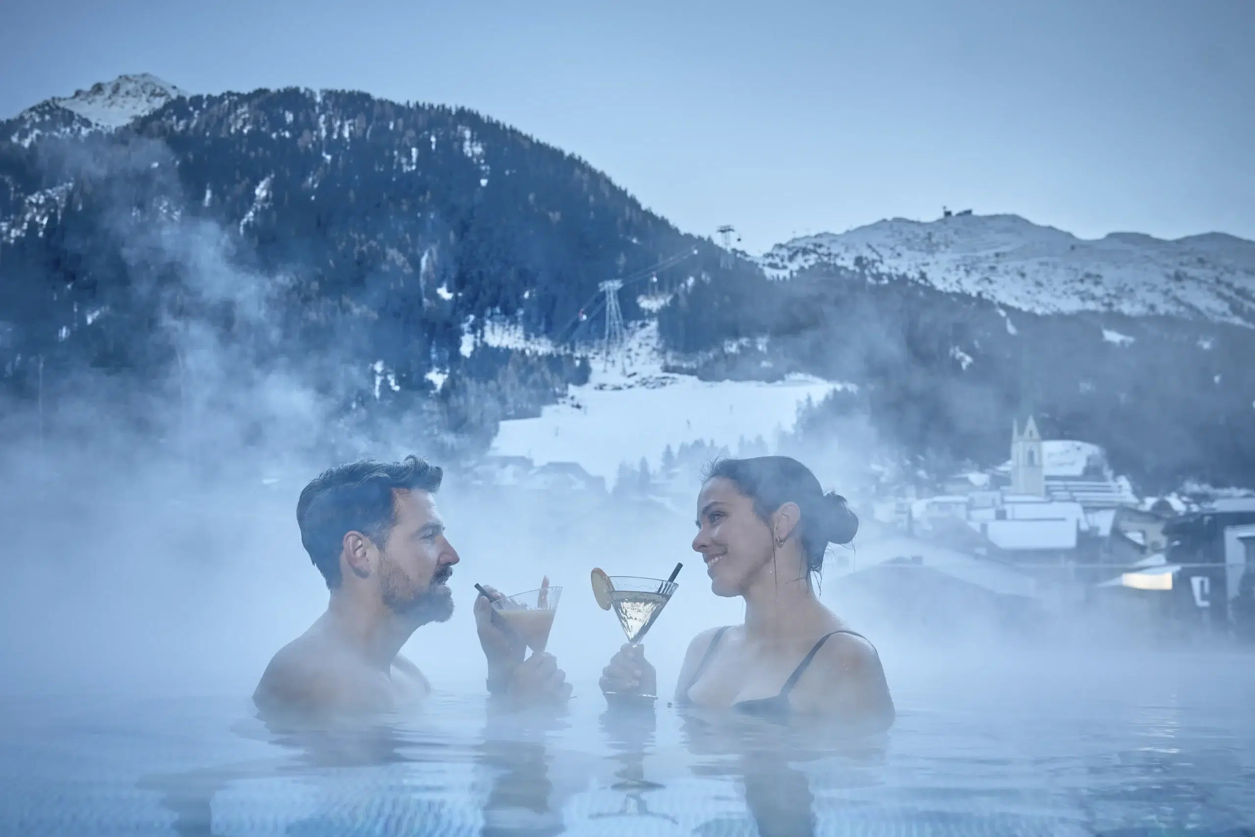 Ein Paar stößt in der Therme eines Wellnesshotels mit Getränken an, im Hintergrund sind schneebedeckte Berge zu sehen.