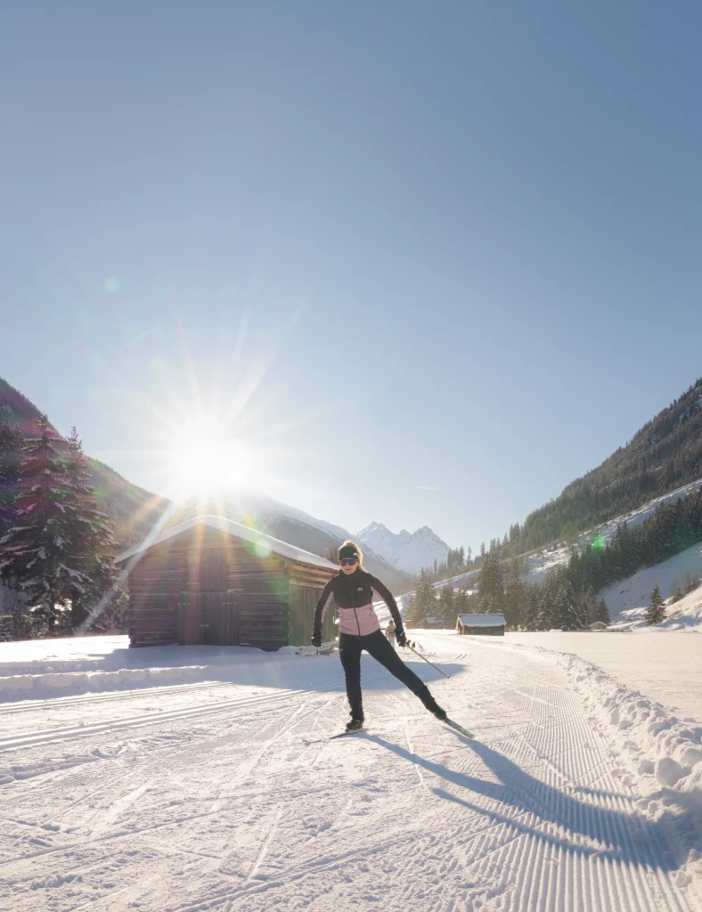 Person, die auf einer verschneiten Winterloipe Ski fährt, mit Bergen und Sonne im Hintergrund.