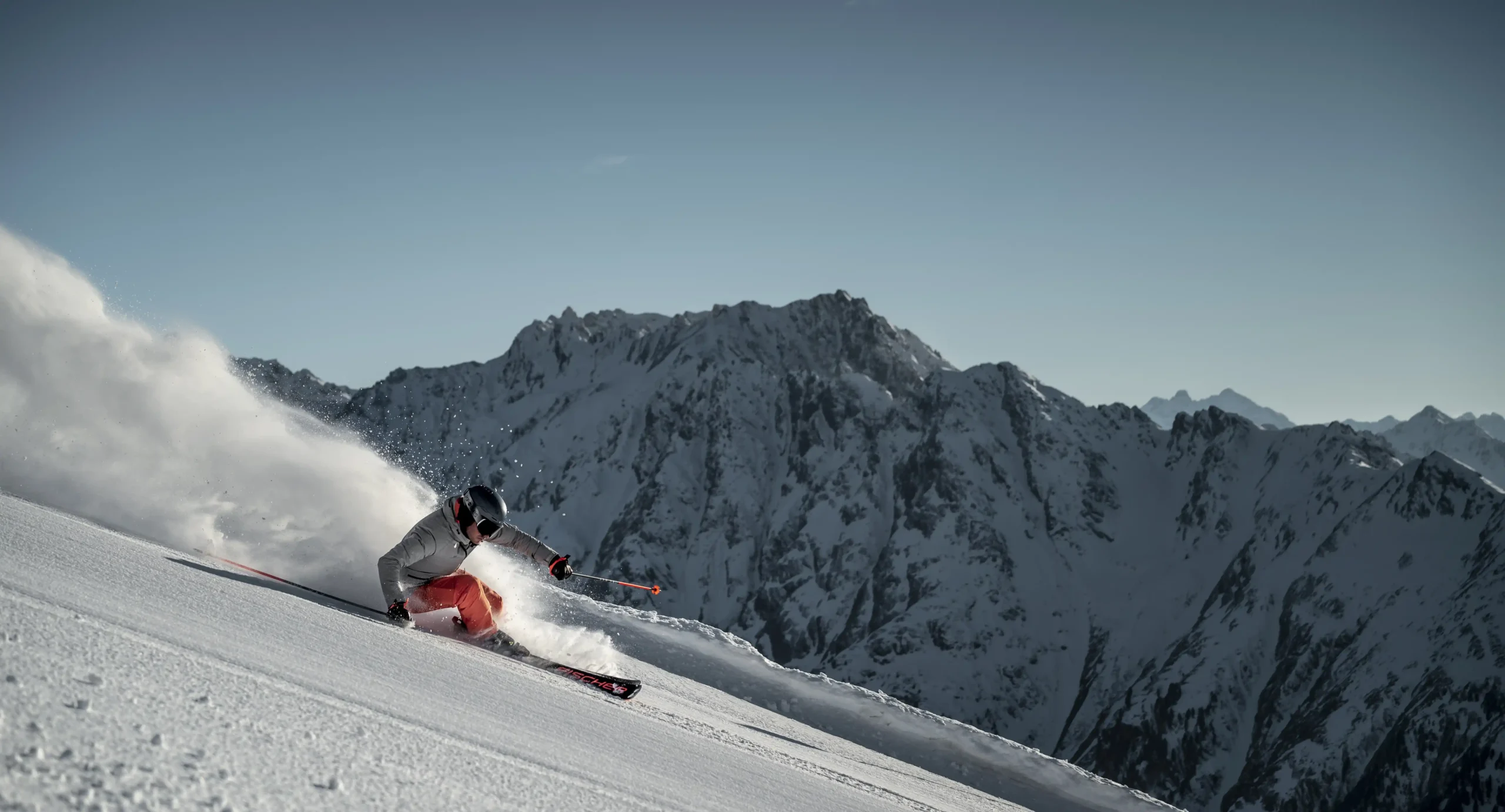 Ein Skifahrer in roten Hosen macht eine scharfe Winterkurve auf einem schneebedeckten Berghang mit Gipfeln im Hintergrund.