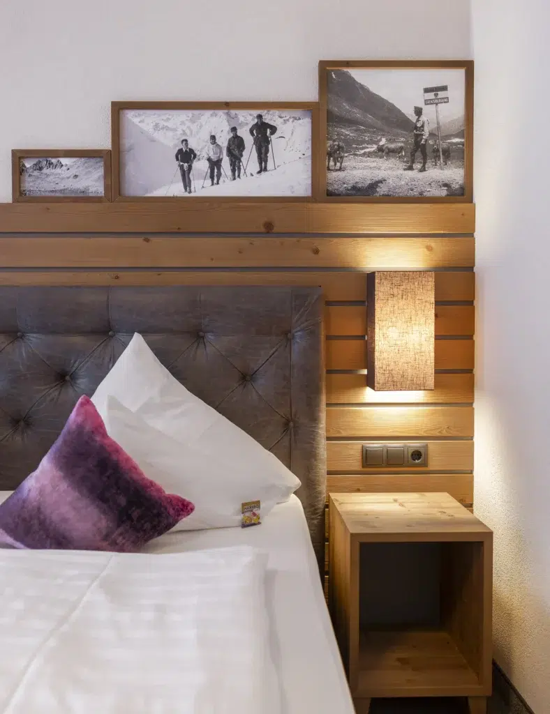 Vista Allegra-Hotelbett mit weißer Bettwäsche, lila Kissen, Holzkopfteil und gerahmten Fotos.