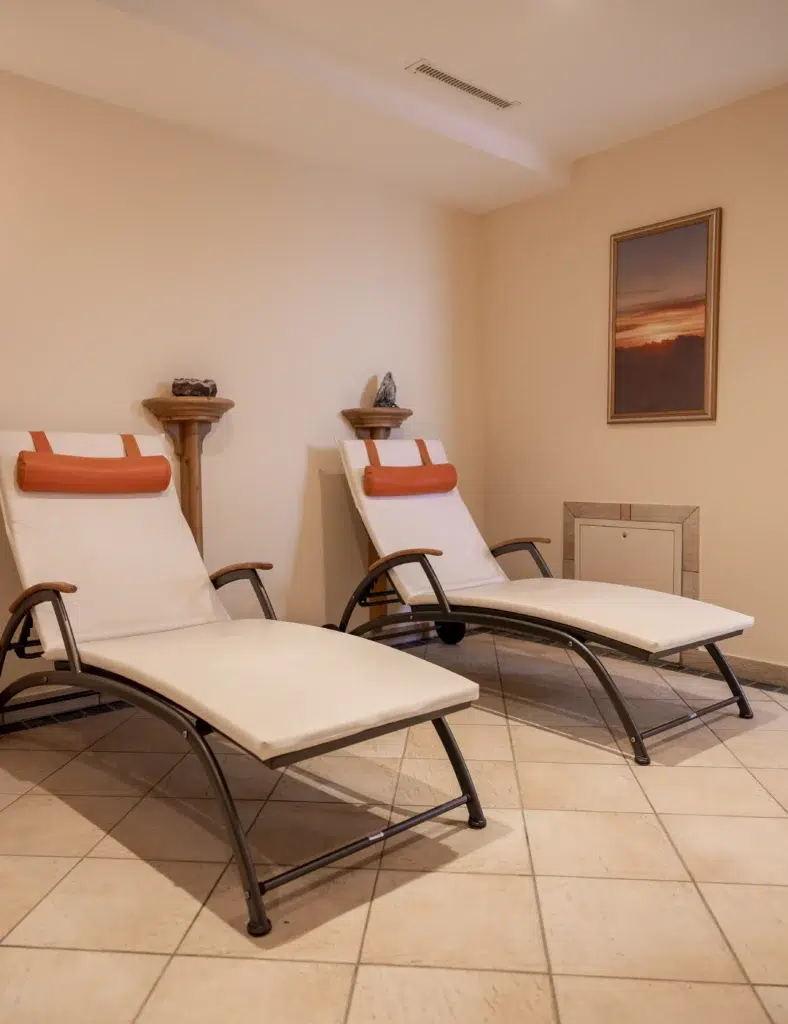 Zwei weiße Loungesessel mit orangefarbenen Kopfstützen in einem beigen Wellnesshotelzimmer mit Wandkunst.