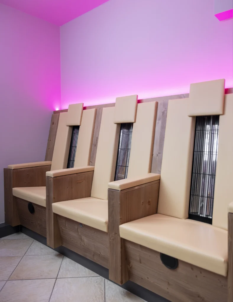 Drei moderne beige Stühle mit Holzarmlehnen in einem Wellnesshotel, unter rosafarbener Ambientebeleuchtung.