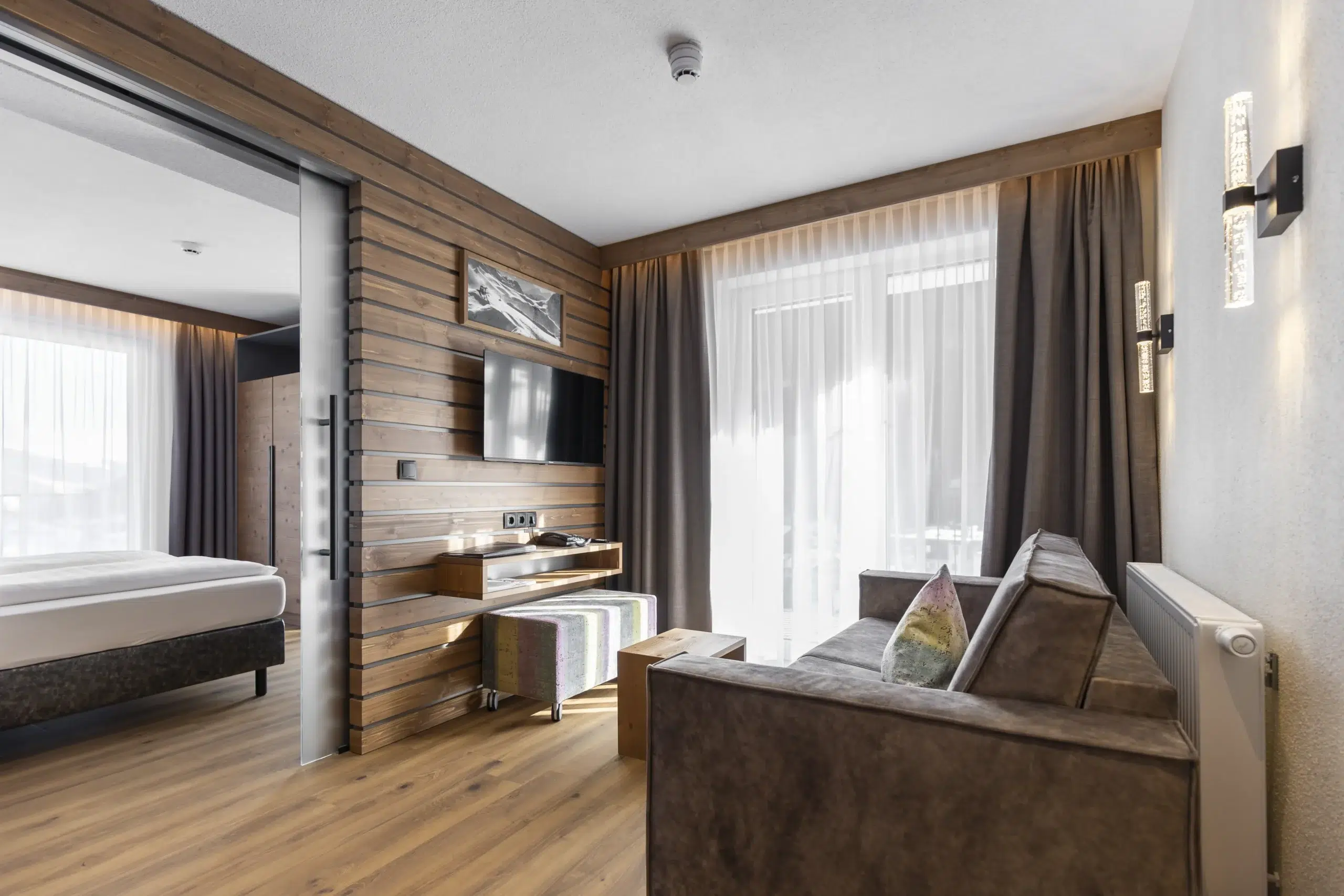 Modernes Vista Allegra-Hotelzimmer mit Bett, Sofa, Fernseher und großen Fenstern mit transparenten Vorhängen.