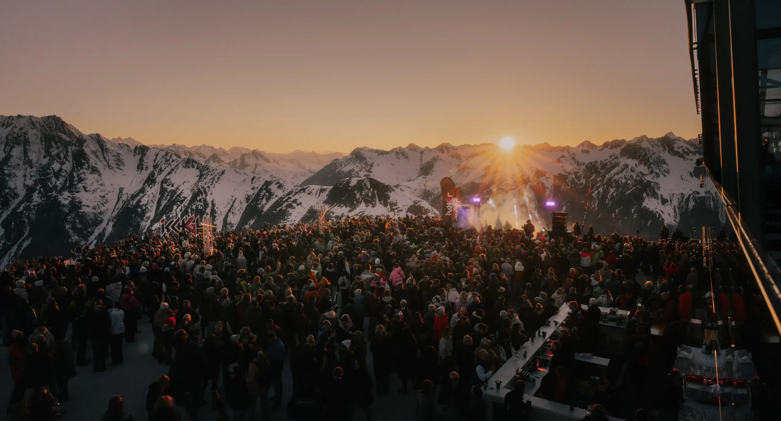 Eine große Menschenmenge im Winter versammelt sich bei Sonnenuntergang auf einem schneebedeckten Berg mit einer Bühne und Lichtern.