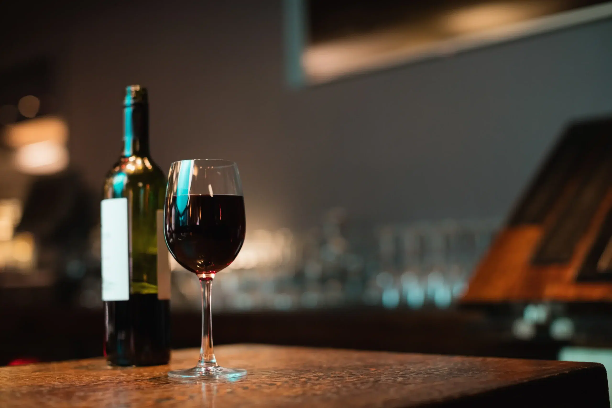 A glass and a bottle of red wine on a wooden restaurant table in a dimly lit setting.