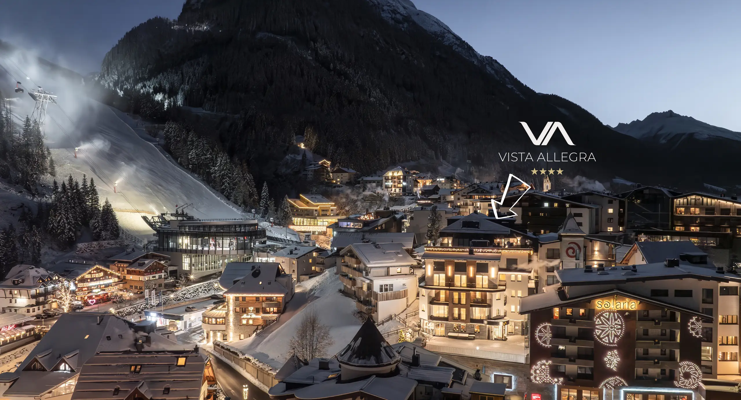 Ein verschneites Bergdorf in der Abenddämmerung, das Vista Allegra-Schild leuchtet zwischen den beleuchteten Gebäuden.