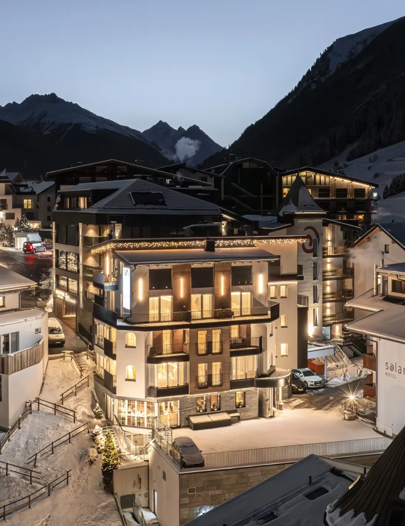 Modernes Hotel Vista Allegra in der Abenddämmerung in einem verschneiten Bergdorf mit Alpengipfeln.